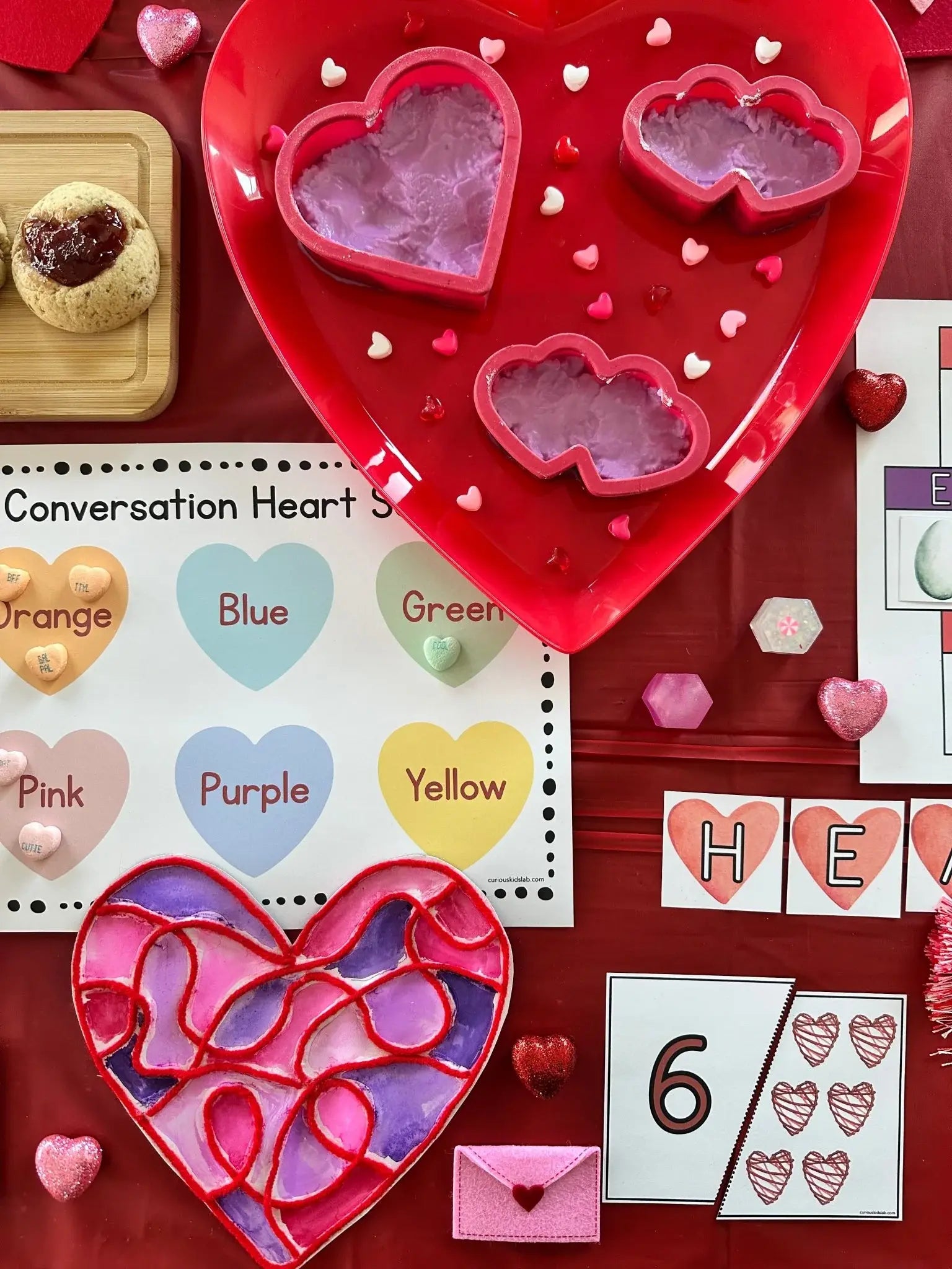Heart-shaped cookie cutters and stained glass hearts on a red surface with educational materials for Valentines Unit Study.