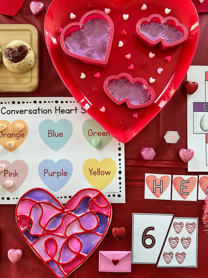 Heart-shaped cookie cutters and stained glass hearts on a red surface with educational materials for Valentines Unit Study.
